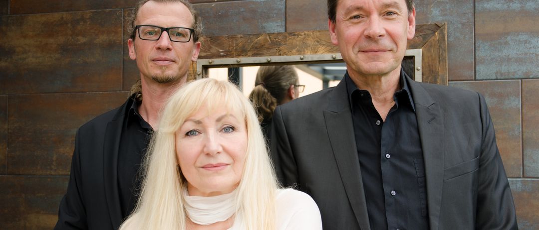 Drei Personen stehen vor einer Metallwand, eine Frau in Weiß und zwei Männer in schwarzen Anzügen. Der Mann links trägt eine Brille.