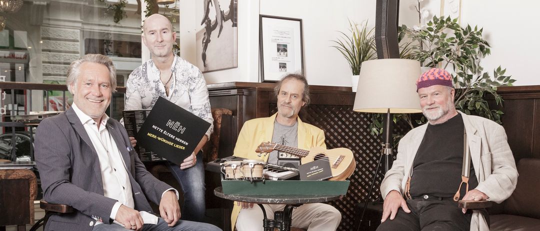 Vier Männer sitzen um einen Tisch mit Gitarren und einem Buch. Sie scheinen für ein Foto in einem Raum mit Holzwänden und einer Lampe zu posieren.