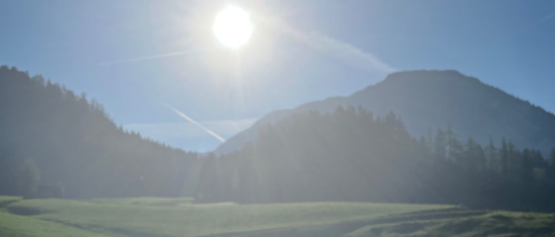 Ein sonniger Tag mit einem klaren blauen Himmel, einem großen grünen Feld und einer kurvenreichen Straße, die hindurchführt. In der Ferne ist ein Gebirgszug sichtbar.