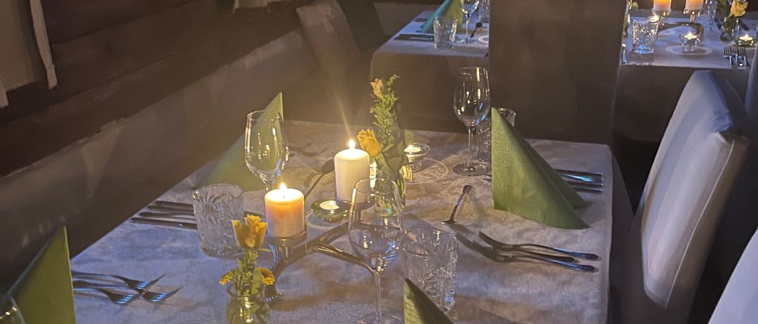 Ein schwach beleuchteter Essbereich mit Holzwänden. Zwei Tische sind mit weißen Tischdecken, Besteck und Kerzen gedeckt. Jeder Tisch hat einen Blumenstrauß als Mittelpunkt. Stühle umgeben die Tische.