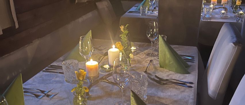 Ein schwach beleuchteter Essbereich mit Holzwänden. Zwei Tische sind mit weißen Tischdecken, Besteck und Kerzen gedeckt. Jeder Tisch hat einen Blumenstrauß als Mittelpunkt. Stühle umgeben die Tische.