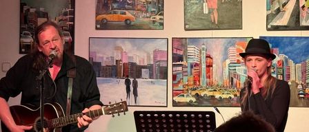 Ein Mann spielt Gitarre vor einer Wand mit Stadtgemälden. Die Bilder zeigen städtische Szenen und Menschen. Der Musiker ist in Schwarz gekleidet.