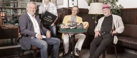 Vier Männer in Anzügen sitzen um einen Tisch in einem Raum, halten Gitarren und lächeln. Einer hält ein Buch mit dem Titel Nettelerster Morgen.