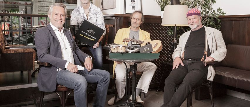 Vier Männer in Anzügen sitzen um einen Tisch in einem Raum, halten Gitarren und lächeln. Einer hält ein Buch mit dem Titel Nettelerster Morgen.