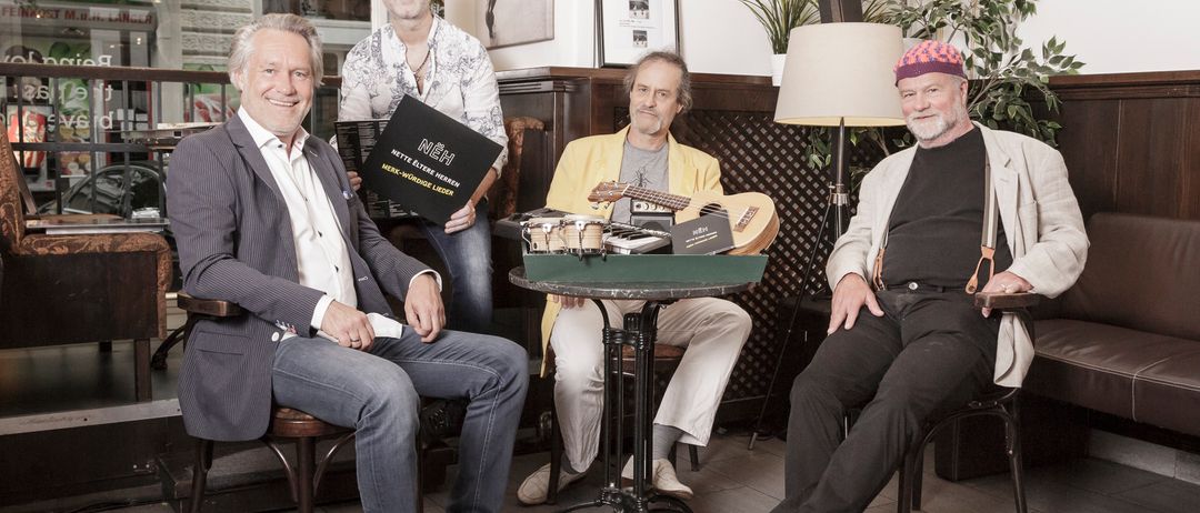 Vier Männer in Anzügen sitzen um einen Tisch in einem Raum, halten Gitarren und lächeln. Einer hält ein Buch mit dem Titel Nettelerster Morgen.