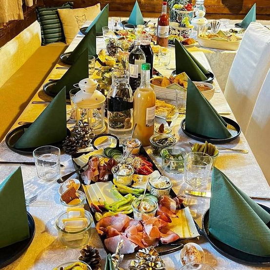 Ein langer Tisch ist mit verschiedenen Speisen wie Fleisch, Käse, Gemüse und Gewürzen gedeckt. Grüne Servietten sind gefaltet und auf schwarzen Tellern platziert. Flaschen mit Saucen und eine Laterne stehen auf dem Tisch.