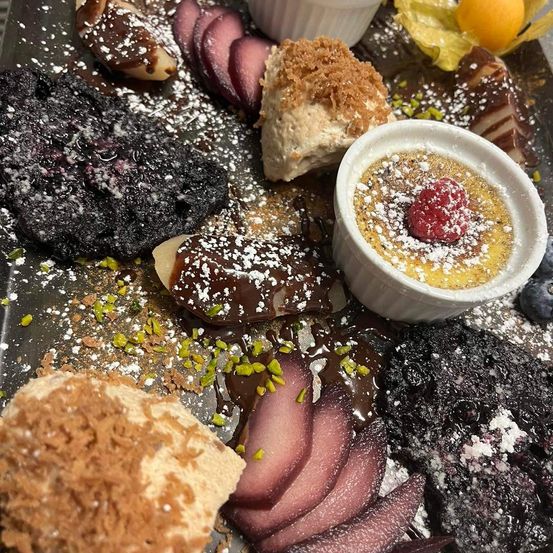 Eine Auswahl an Desserts ist auf einem Tablett angerichtet, darunter eine Creme Brulee mit einer Himbeere, schokoladenüberzogene Gebäckstücke und Obstscheiben.