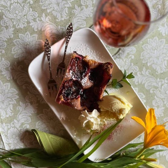 Ein Stück Kirschtarte mit Eiscreme und einem Glas Wein auf einer Tischdecke mit Blumenmuster.