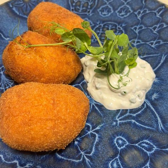 Drei goldbraune Kroketten auf einem blauen Blumenteller mit einer cremigen Dip-Sauce und grüner Garnierung.