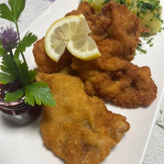 Ein weißer Teller mit gebratenem Fisch, garniert mit Zitronenscheiben und Petersilie, begleitet von einem kleinen Glas Marmelade und einer Beilage aus grünem Gemüse.