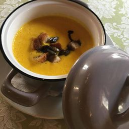 Eine Schüssel mit cremiger Kürbissuppe, garniert mit gerösteten Brotwürfeln und Kürbiskernen auf einem Tisch mit Blumenmuster.