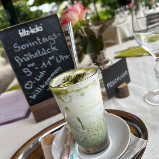Ein grünes Getränk in einem hohen Glas mit Strohhalm, auf einem silbernen Teller auf einem Tisch, mit einem Kreidetafelschild im Hintergrund.
