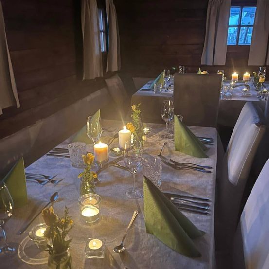 Ein warm beleuchteter Essbereich mit Holzwänden, der mit Kerzen, Blumen und Besteck gedeckte Tische aufweist. Der Raum hat eine gemütliche Atmosphäre.