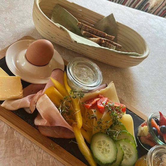 Ein Frühstücksbrett mit verschiedenen Fleischsorten, Käse, Eiern und Gemüse auf einem Holzbrett, begleitet von einem Korb mit Brot und einer Schüssel mit Obst.