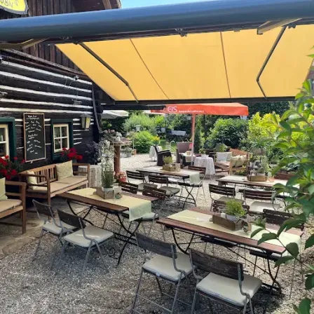 Ein Restaurant im Freien mit einer Holzbank, einem Tisch mit einem Topf und einem gelben Baldachin. Das Restaurant hat einen Garten mit Pflanzen und Blumen.