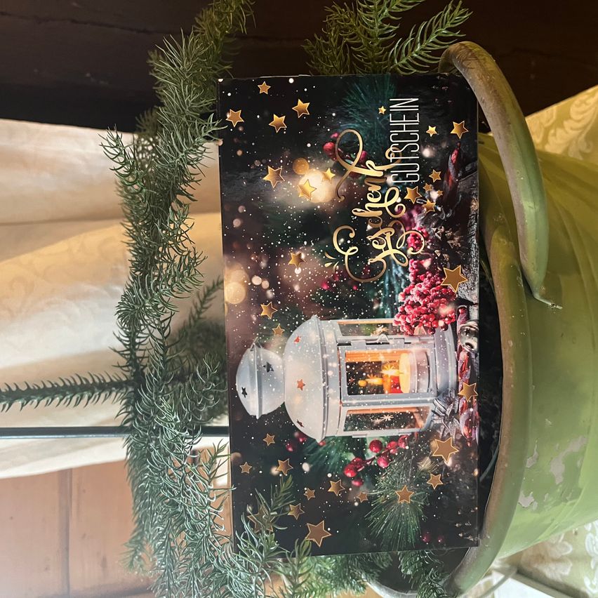 Eine Weihnachtskarte mit einer Laterne und Sternen, umgeben von festlicher Grünpflanze, liegt auf einem grünen Pflanzentopf.