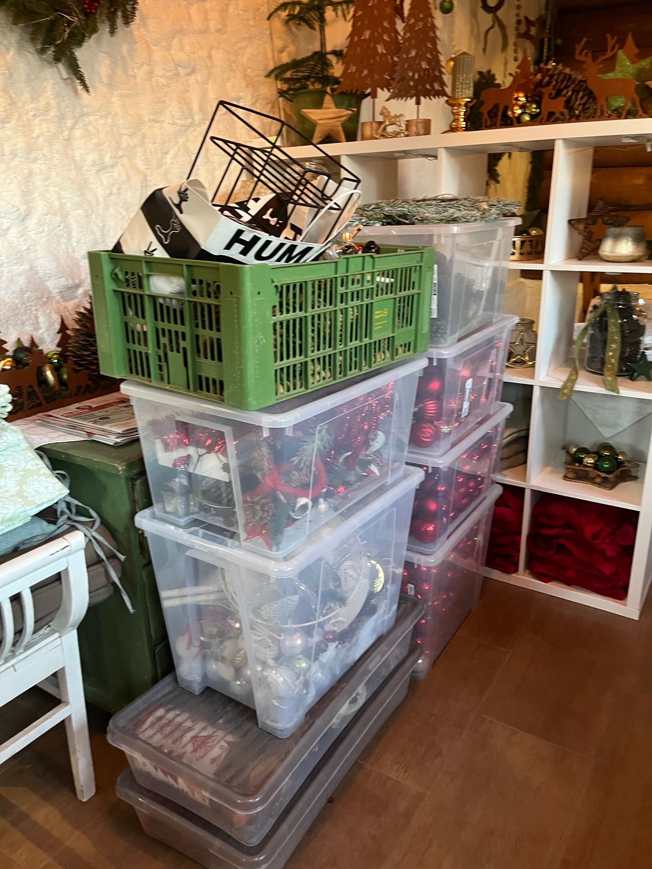 A room filled with storage boxes and crates, containing Christmas decorations. One green crate with 'HUM' on it sits atop others. Shelves in the background hold more festive items.