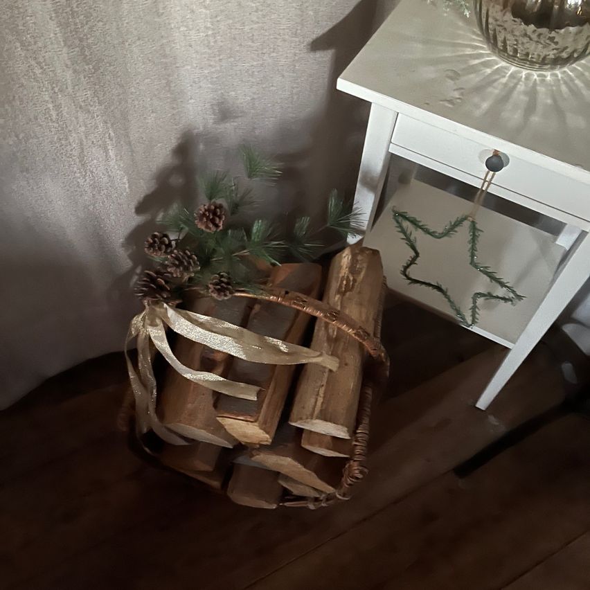 A wicker basket filled with firewood, adorned with pine cones, is placed next to a white table with a silver ornament on top.
