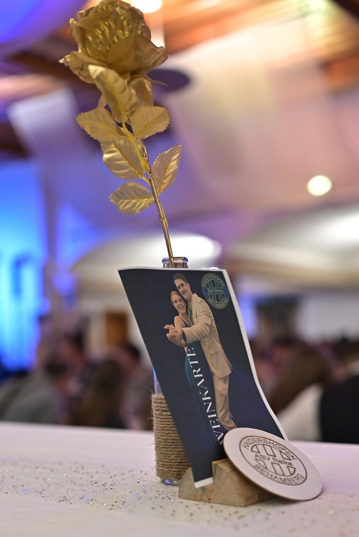 Ein Ausschnitt einer Hochzeitseinladung wird gezeigt, mit einem goldenen Blatt als Mittelpunkt in einer Glasflasche. Im Hintergrund ist eine verschwommene Gruppe von Menschen zu sehen.