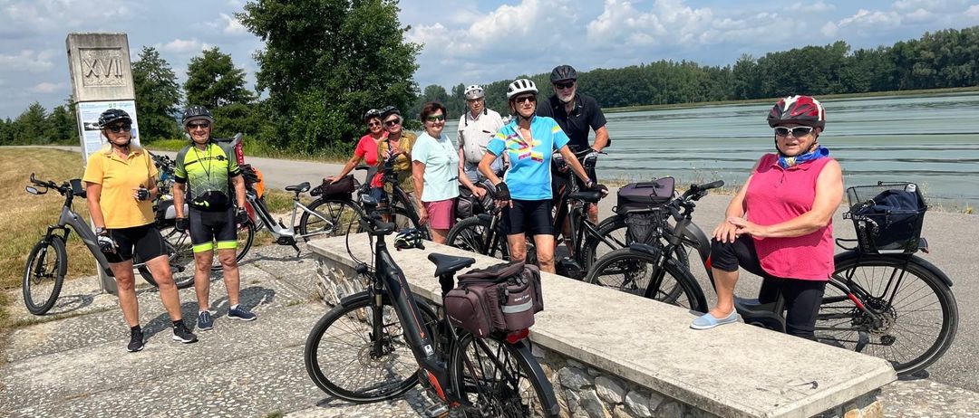 Eine Gruppe von Radfahrern, alle mit Helmen und Fahrradausrüstung, posiert für ein Foto am See. Sie stehen und sitzen auf ihren Fahrrädern, einige lehnen sich auf einer Steinbank aus.