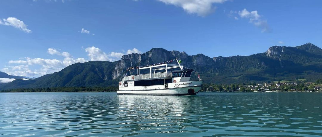 Ein weißes Boot namens McMondseeland fährt auf einem ruhigen See mit Bergen im Hintergrund unter einem blauen Himmel mit Wolken.