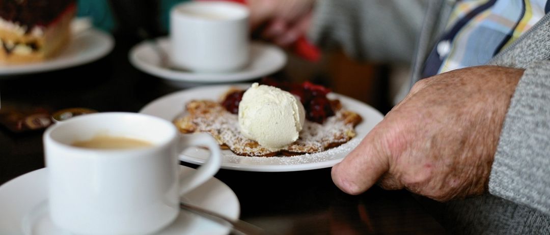 Ein Nahaufnahme eines Tisches mit zwei weißen Tassen, eine mit Kaffee und die andere mit Eiscreme auf einem Dessert. Eine Hand hält ein rotes Taschentuch.