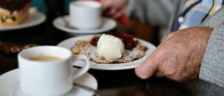 Ein Nahaufnahme eines Tisches mit zwei weißen Tassen, eine mit Kaffee und die andere mit Eiscreme auf einem Dessert. Eine Hand hält ein rotes Taschentuch.