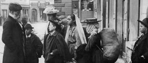 Ein Schwarz-Weiß-Foto einer Gruppe von Menschen, die auf einem Bürgersteig stehen und teilweise Gepäck tragen. Sie scheinen auf etwas zu warten. Der Hintergrund zeigt eine urbane Umgebung mit Gebäuden und einem Schild.
