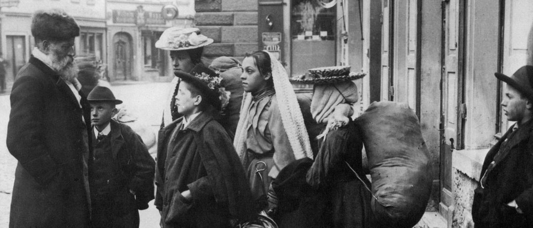 Ein Schwarz-Weiß-Foto einer Gruppe von Menschen, die auf einem Bürgersteig stehen und teilweise Gepäck tragen. Sie scheinen auf etwas zu warten. Der Hintergrund zeigt eine urbane Umgebung mit Gebäuden und einem Schild.