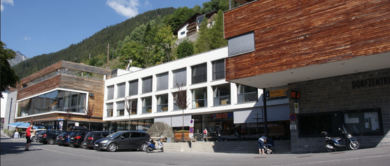 Ein modernes Gebäude mit Holzfassade steht an einer Straße mit geparkten Autos und einer Person mit Kinderwagen davor. Ein Berg ist im Hintergrund.