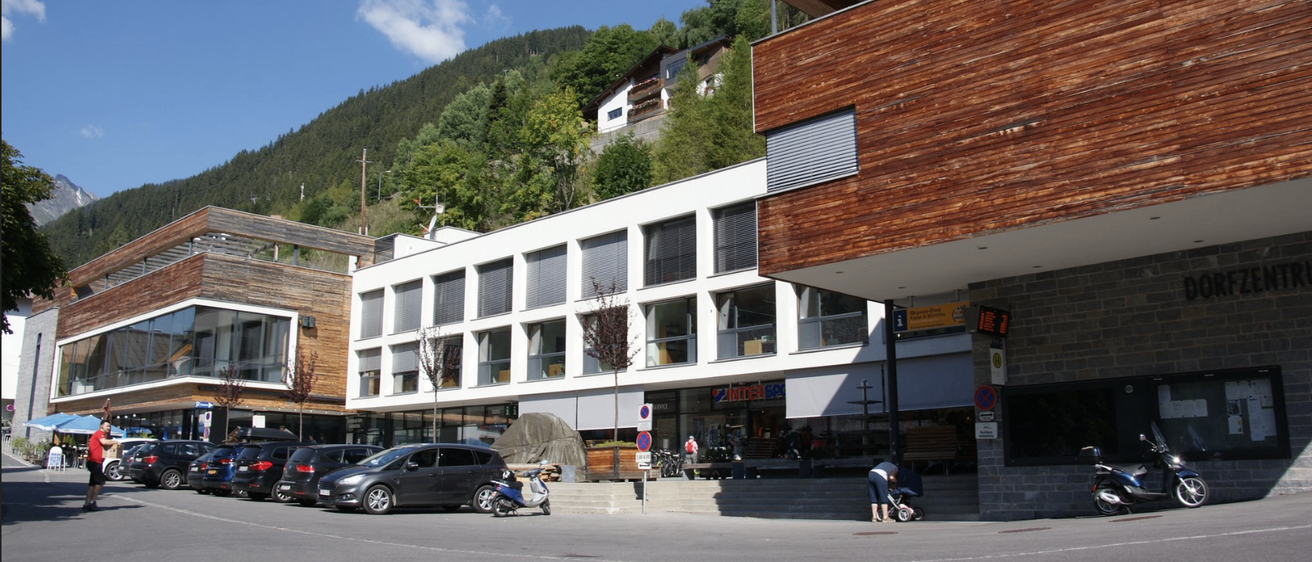 Ein modernes Gebäude mit Holzfassade steht an einer Straße mit geparkten Autos und einer Person mit Kinderwagen davor. Ein Berg ist im Hintergrund.