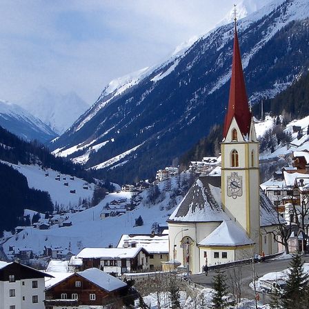 Ein kleines Dorf mit einer Kirche im Vordergrund, umgeben von schneebedeckten Bergen und Gebäuden.