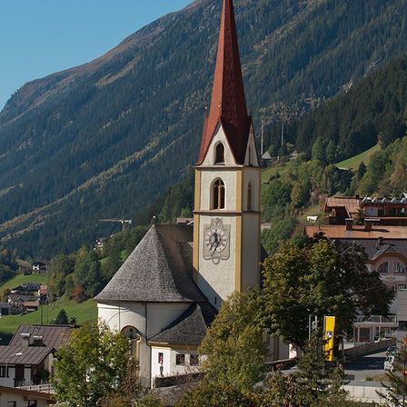 Eine Kirche mit Turm und Uhr steht in einem Bergdorf mit üppigen grünen Hügeln und dichtem Wald im Hintergrund.