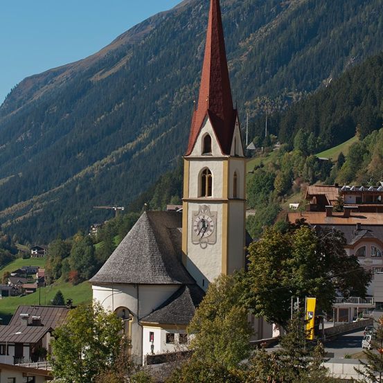 Eine Kirche mit Turm und Uhr steht in einem Bergdorf mit üppigen grünen Hügeln und dichtem Wald im Hintergrund.