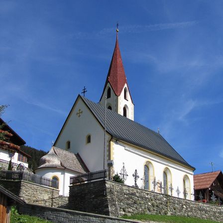 Eine kleine weiße Kirche mit einem roten Turm und einem schwarzen Dach steht auf einem Hügel unter einem klaren blauen Himmel.