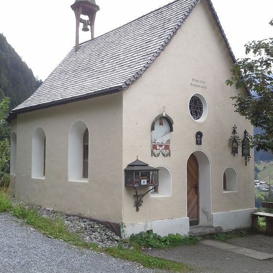 Eine kleine Kapelle mit einem Glockenturm, einer Holztür und einem Fenster mit einem Buntglasdesign. Die Kapelle hat ein graues Dach und ist von Gras und Steinen umgeben.
