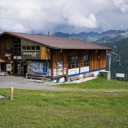 Ein holzverkleidetes Gebäude mit braunem Dach und blauem Rand steht am Berghang, mit einem Schild, das 'DIASBAHN 1830m' anzeigt. Bänke und ein grüner Container sind in der Nähe, und Berge sind im Hintergrund.