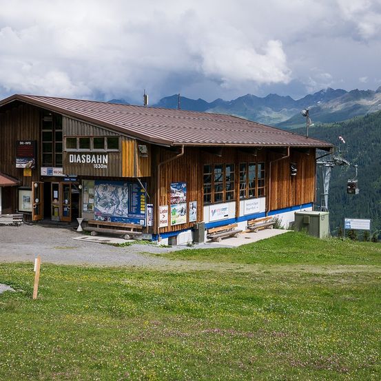 Ein holzverkleidetes Gebäude mit braunem Dach und blauem Rand steht am Berghang, mit einem Schild, das 'DIASBAHN 1830m' anzeigt. Bänke und ein grüner Container sind in der Nähe, und Berge sind im Hintergrund.