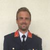 Ein Mann in einem dunkelblauen Uniform mit goldenen Knöpfen, einem roten Stern und einem Abzeichen lächelt gegen eine beige Wand in die Kamera.
