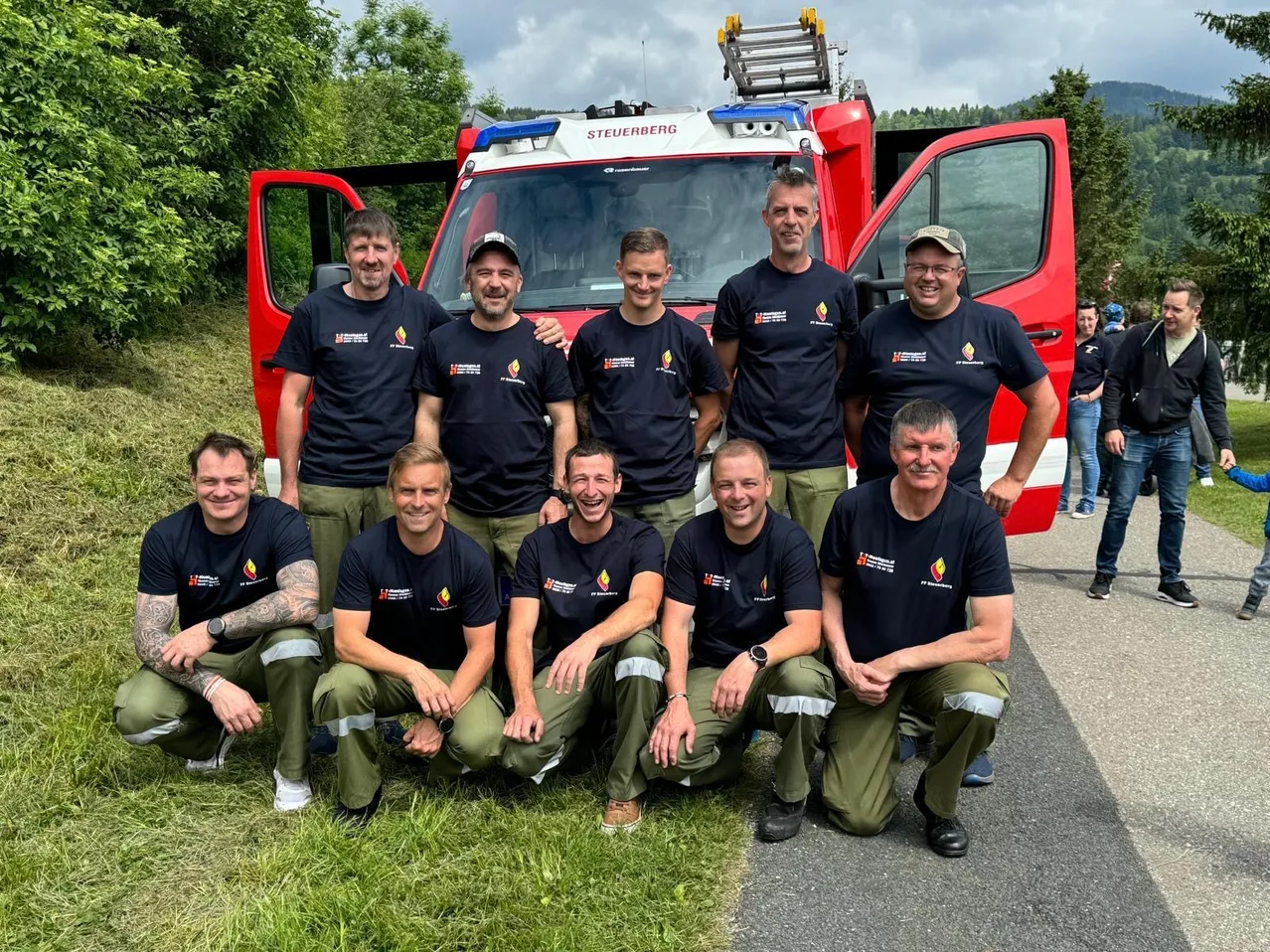 Eine Gruppe Feuerwehrleute posiert für ein Foto vor einem roten Feuerwehrauto mit offenen Türen, mit grünem Gras und Bäumen im Hintergrund.