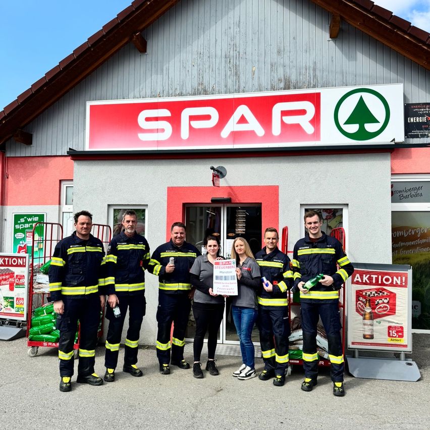 Eine Gruppe von Feuerwehrleuten steht vor einem Spar-Laden und hält einen Scheck, eine Frau steht in der Mitte.