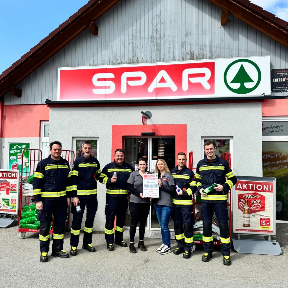Eine Gruppe von Feuerwehrleuten steht vor einem Spar-Laden und hält einen Scheck, eine Frau steht in der Mitte.