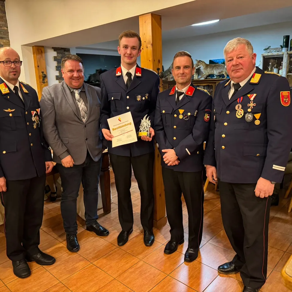 Fünf Männer in Uniform posieren für ein Foto. Einer hält eine Auszeichnung und ein Zertifikat. Der Raum hat geflieste Böden und Holzsäulen.