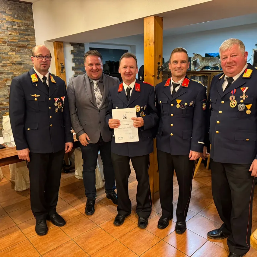Fünf Männer in Militäruniformen posieren für ein Foto. Einer hält eine Urkunde. Sie stehen drinnen auf einem Holzboden.