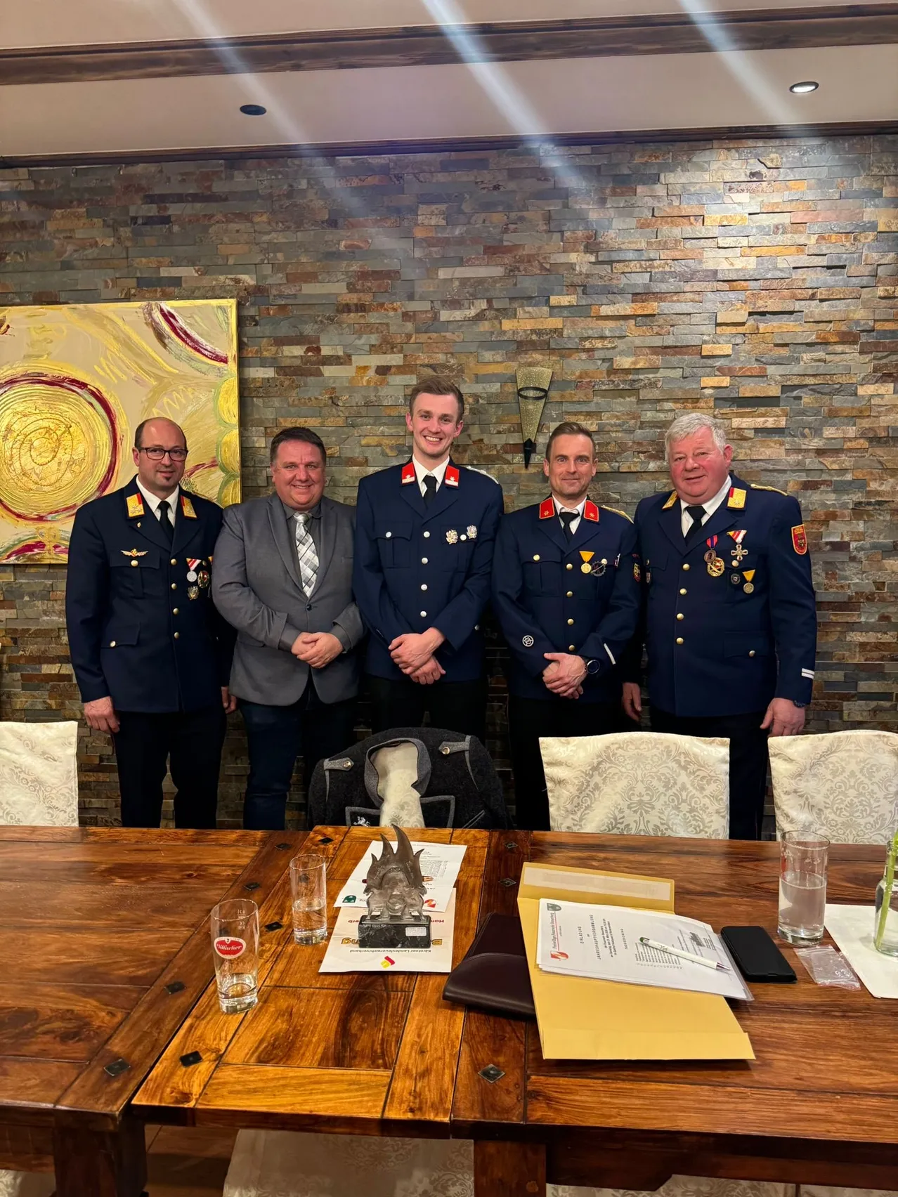 Fünf Männer in Uniform stehen lächelnd für ein Foto hinter einem Tisch. Ein Mantel hängt auf dem Stuhl hinter ihnen, und Papiere und ein Pokal sind auf dem Tisch.