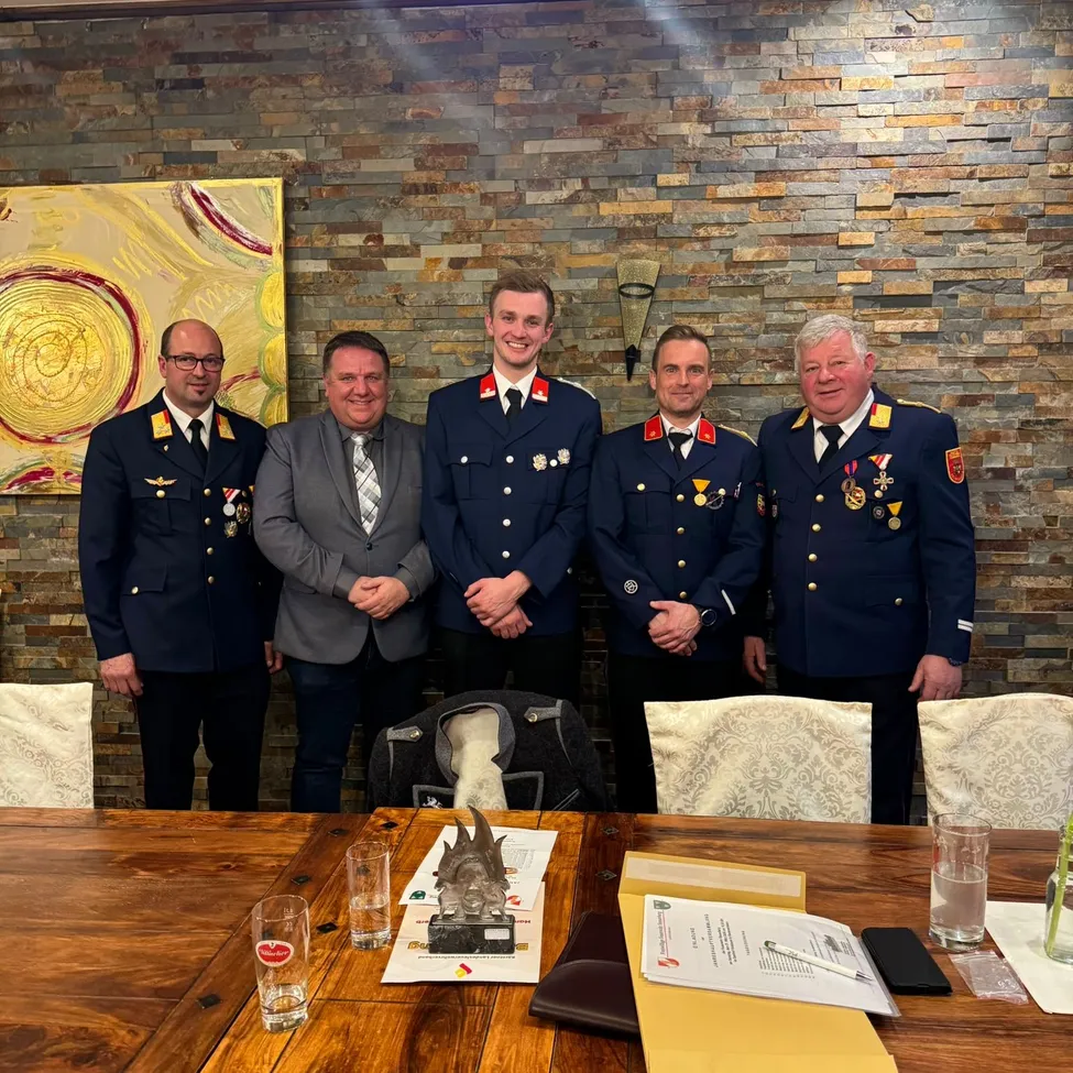 Fünf Männer in Uniform stehen lächelnd für ein Foto hinter einem Tisch. Ein Mantel hängt auf dem Stuhl hinter ihnen, und Papiere und ein Pokal sind auf dem Tisch.