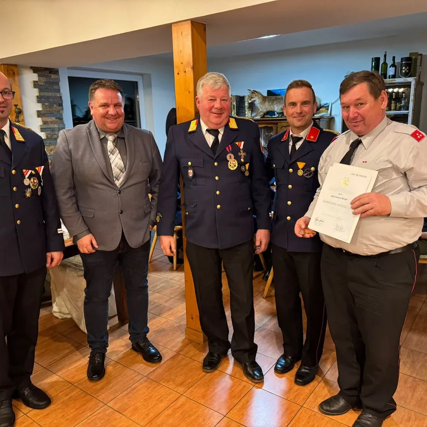 Fünf Männer in Uniformen stehen im Innenraum und lächeln. Einer hält ein Zertifikat und trägt einen grauen Anzug. Ein anderer trägt eine Militäruniform mit Medaillen. Dahinter steht ein Regal mit Flaschen.