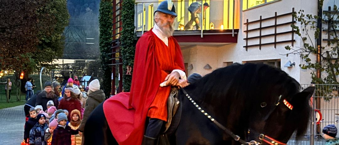 Ein Mann in traditioneller Kleidung reitet auf einem schwarzen Pferd an einem Gebäude vorbei, während Kinder mit Laternen folgen.