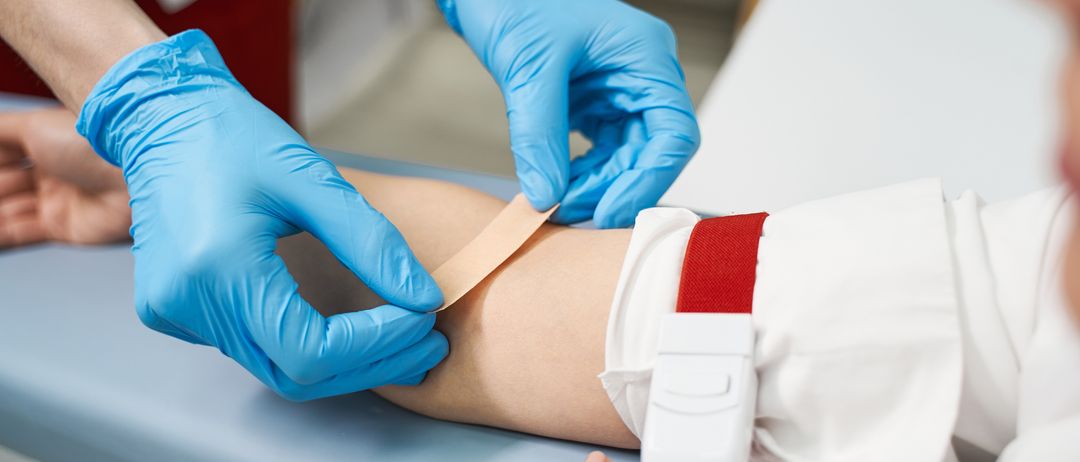 Eine Gesundheitskraft in blauen Handschuhen legt einem Patienten auf einem blauen Tisch einen Verband am Arm an. Der Patient hat einen roten Riemen am Arm.