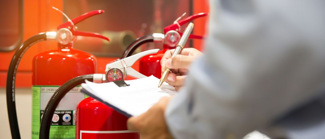 Eine Person in einem grauen Hemd schreibt mit einem Stift auf einem Blatt Papier, während sie vor mehreren Feuerlöschern steht.